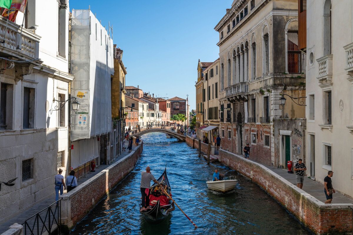 A gondolier rowing a gondola down a quiet canal between old buildings, with a bridge in the distance