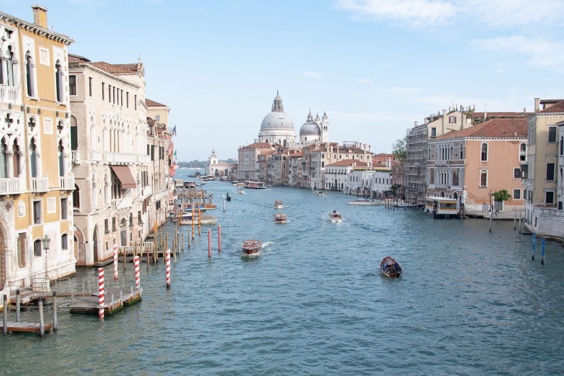 The Grand Canal in Venice on a sunny day, lined with old palaces and striped mooring poles, with motorboats on the water and the domes of Santa Maria della Salute in the distance