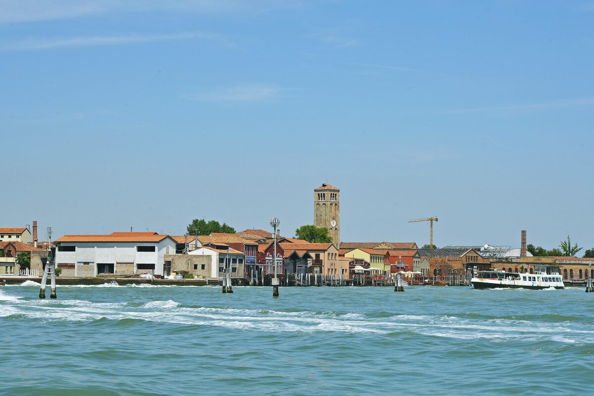 The low waterfront of Murano seen from the lagoon, with colorful houses, a brick bell tower and a vaporetto