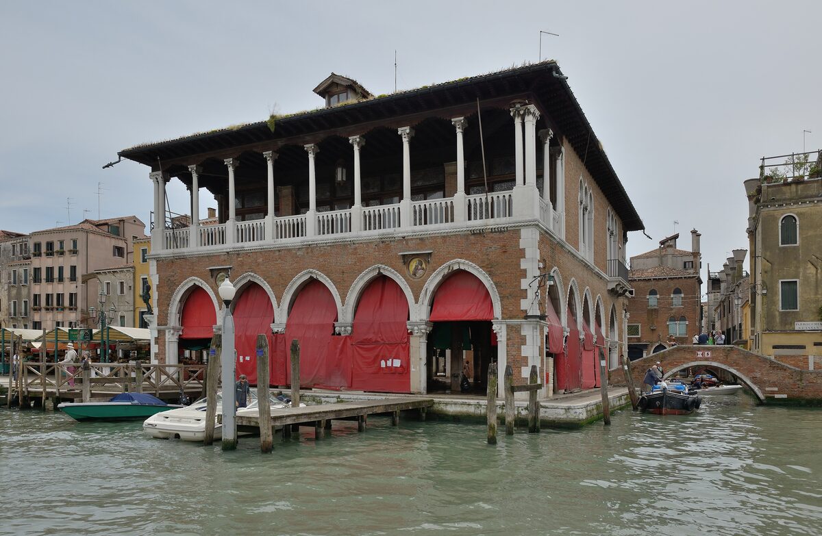 The open, columned hall of the Pescaria fish market at Rialto, with red doors and boats moored on the Grand Canal in front