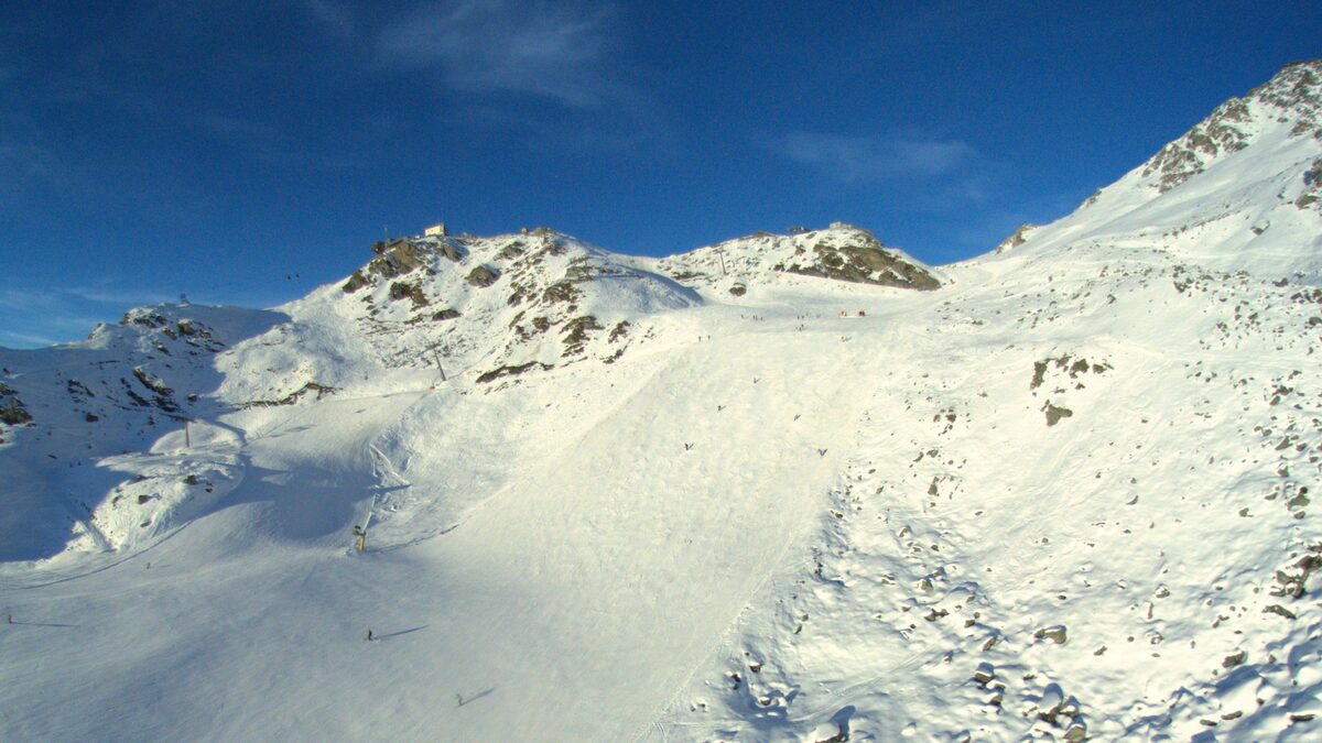 A wide snowy slope with ski tracks and rocky ridges under a blue sky
