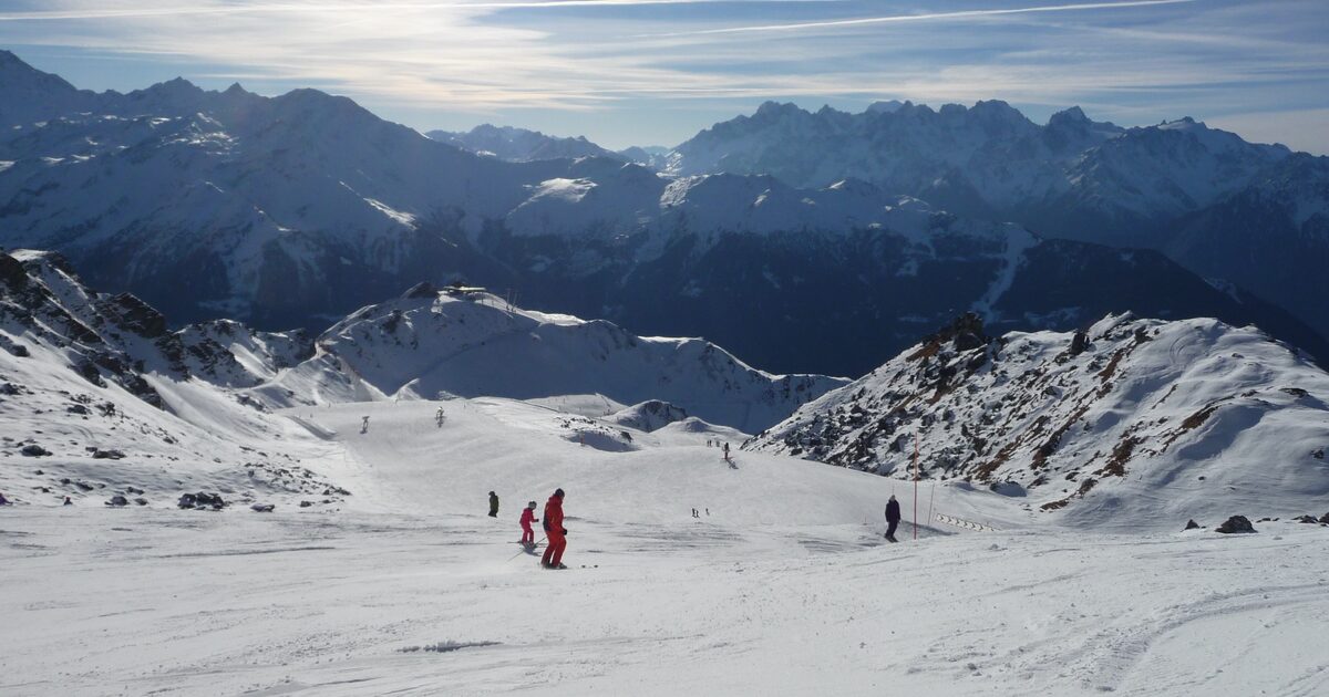 Skiers on a wide sunlit snowy piste, with a row of snow-covered peaks on the horizon