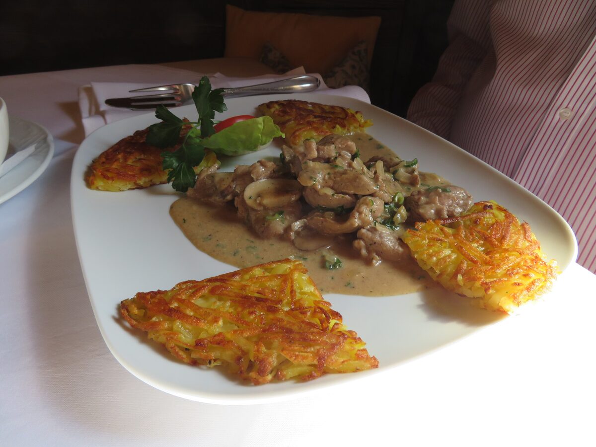 Sliced veal in a cream sauce with mushrooms, next to two golden rösti cakes on a white plate