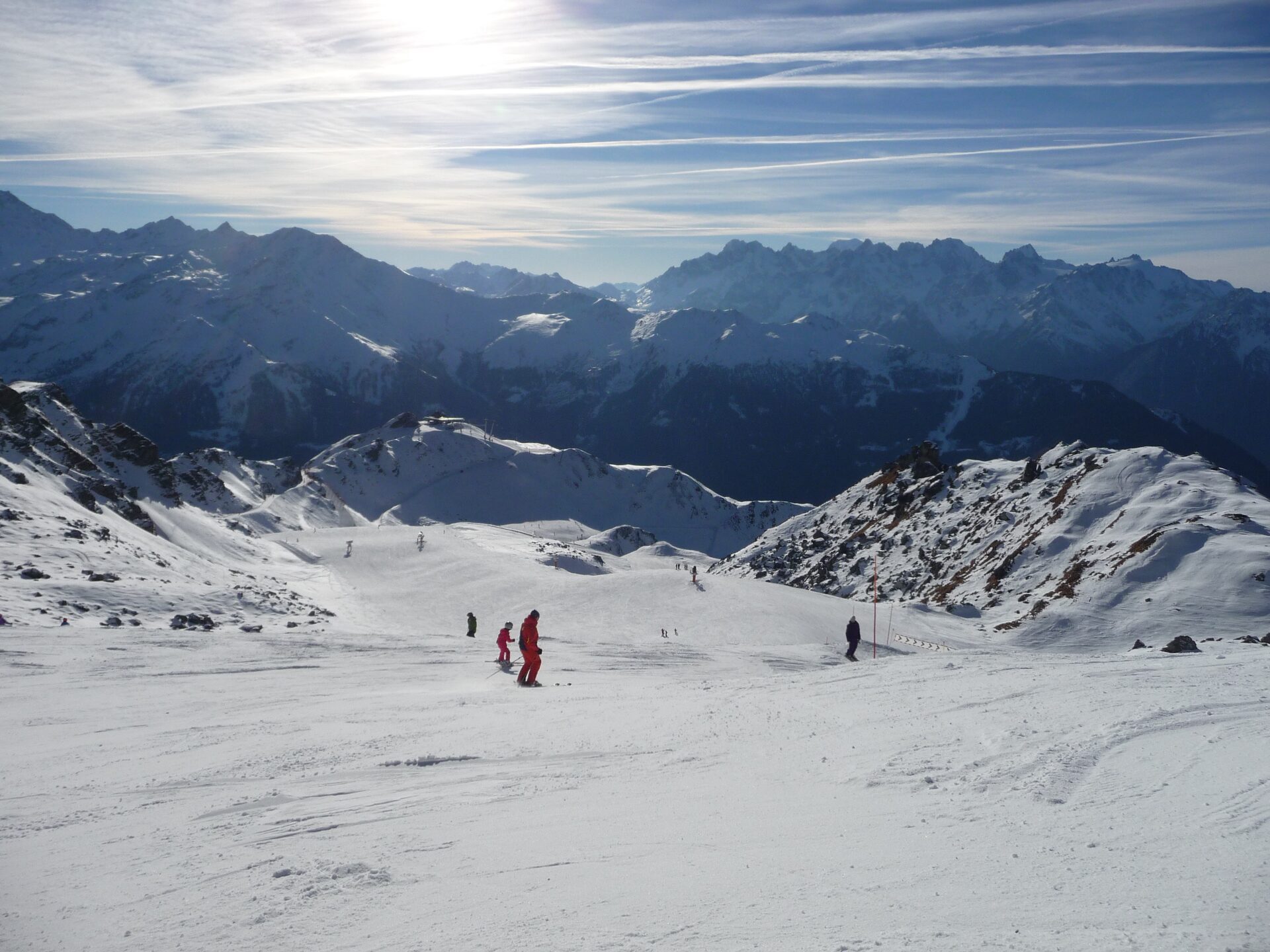 Skiers on a wide sunlit snowy piste, with a row of snow-covered peaks on the horizon