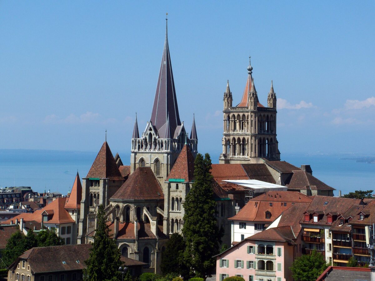 The spire and bell tower of Lausanne cathedral above red-tiled roofs, with Lake Geneva behind under a blue sky