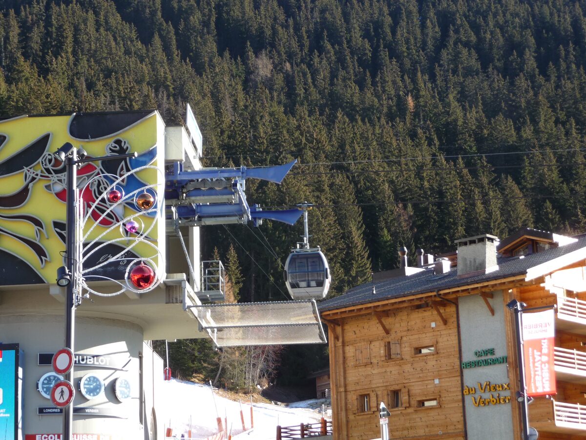 A gondola cabin leaving a station building in Verbier, beside a wooden chalet and a forested slope