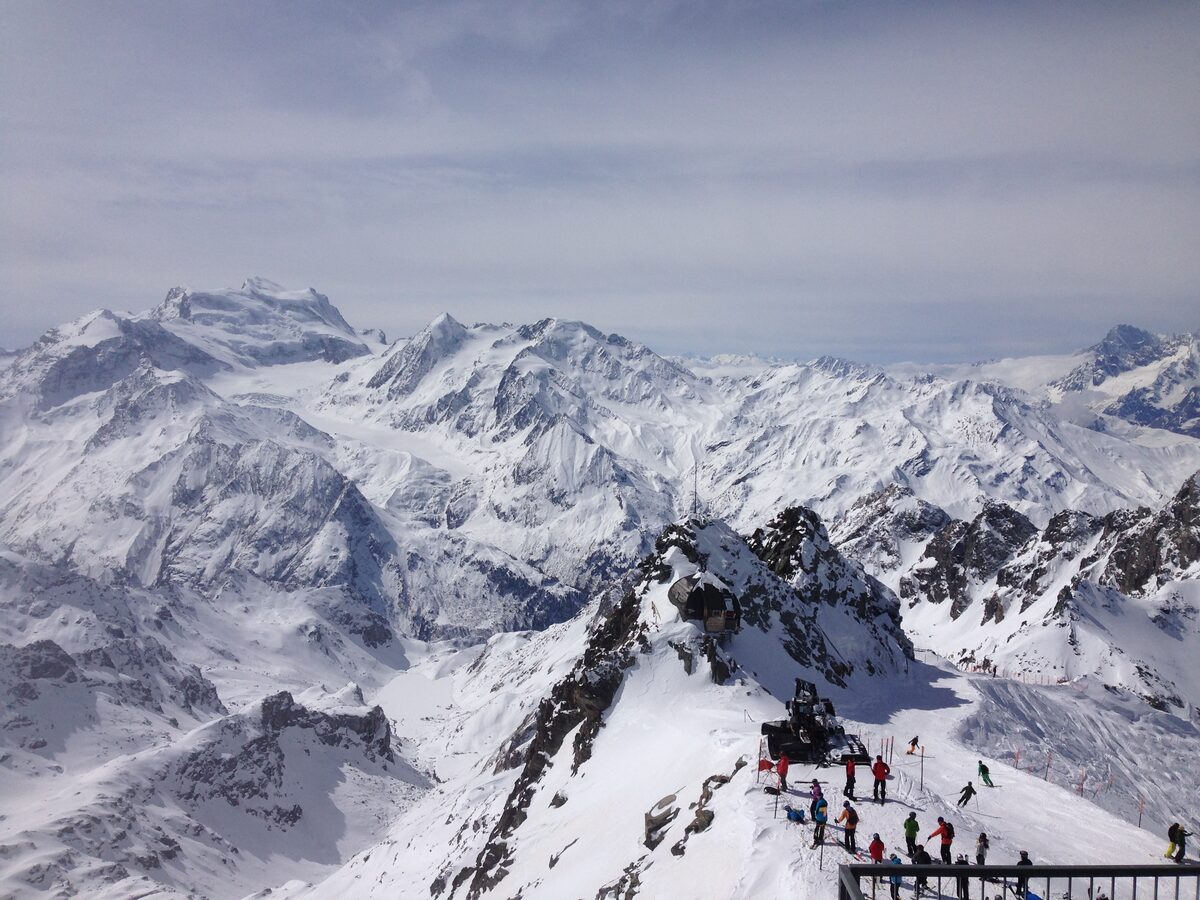 Skiers on the viewing platform of a rocky summit, with a wide range of snow-covered peaks and glaciers below