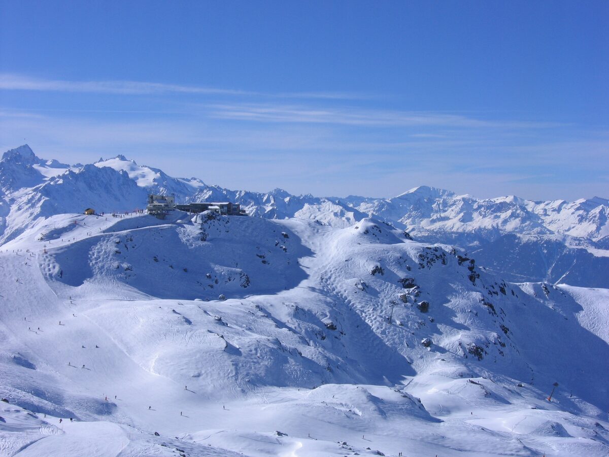 A lift station on a snowy ridge above wide slopes dotted with tiny skiers, with a range of peaks beyond