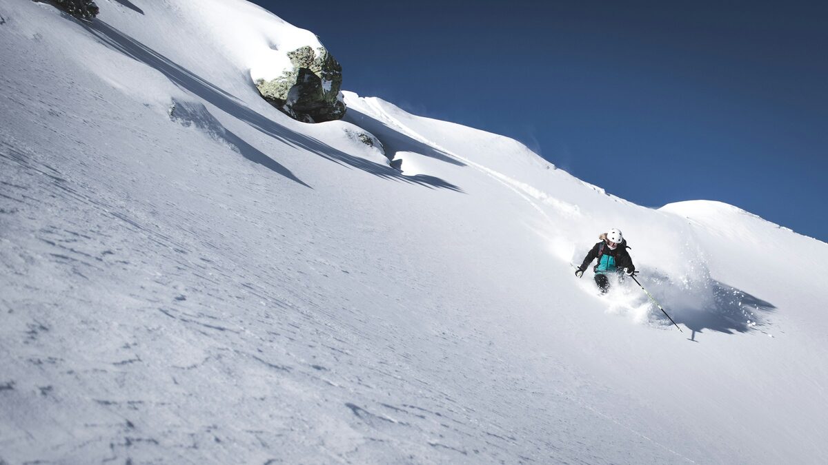 A skier carving through deep powder on a steep snowy slope under a blue sky