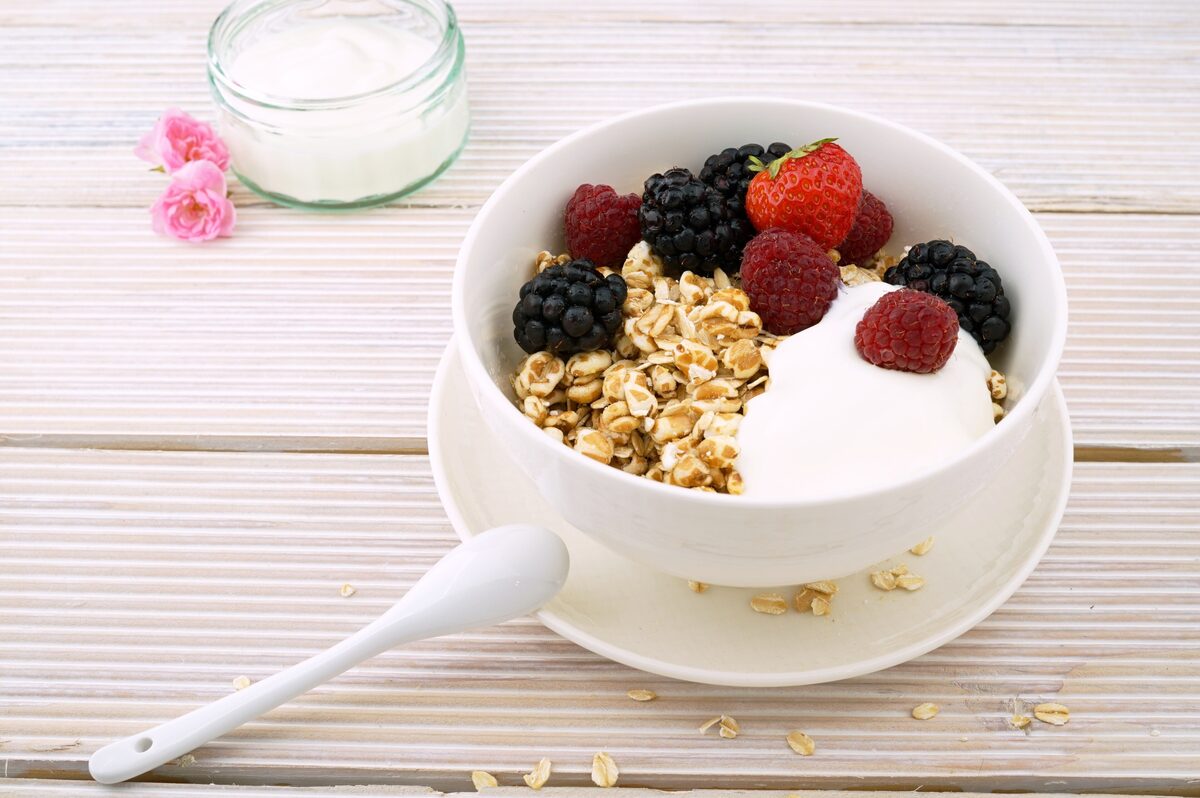 A white bowl of muesli with a scoop of yogurt, blackberries, raspberries and a strawberry on a pale wooden table