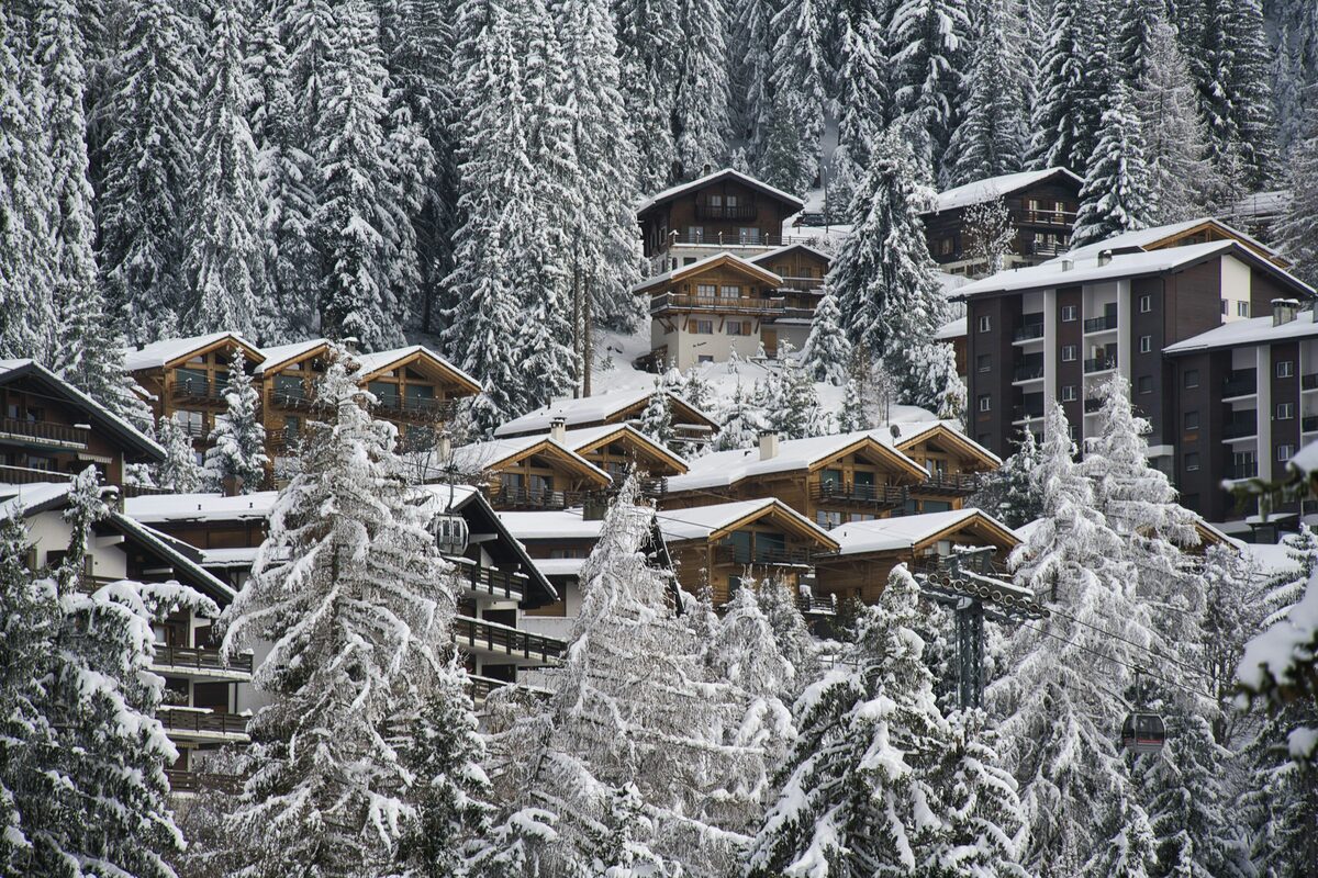 Wooden chalets and apartment blocks among snow-covered fir trees on a hillside, with a gondola cabin on its cable at the lower right