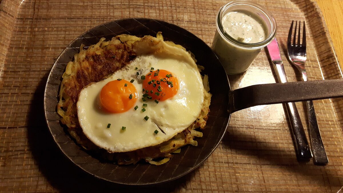A rösti in an iron pan topped with two fried eggs and chives, with a small glass of sauce beside it