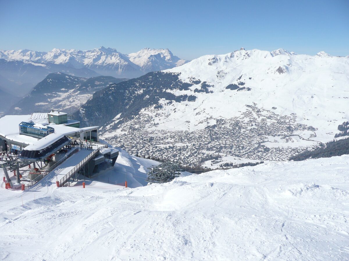 The Les Ruinettes lift station on a snowy ridge, with the village of Verbier in the valley and snow-covered mountains beyond