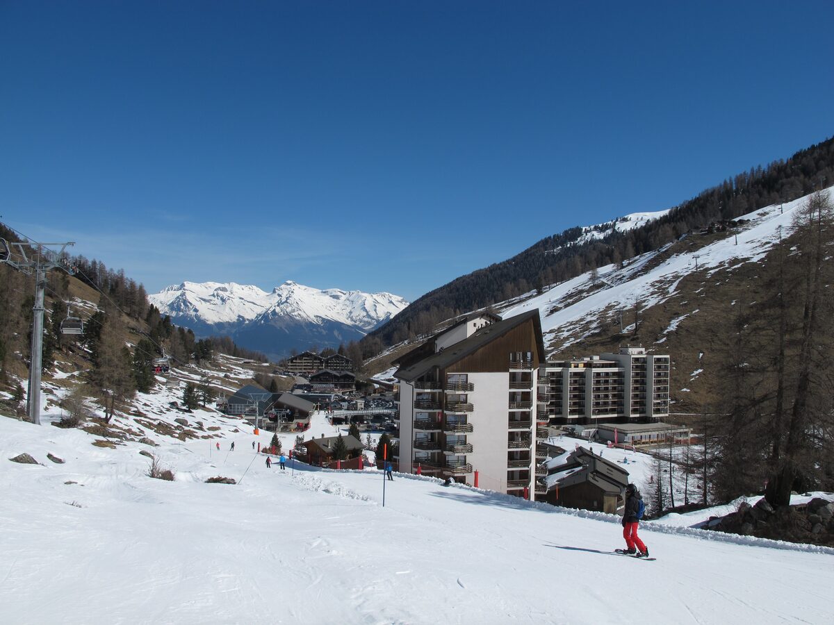 A ski slope in front of apartment blocks, with a snowboarder in red trousers and snowy mountains behind