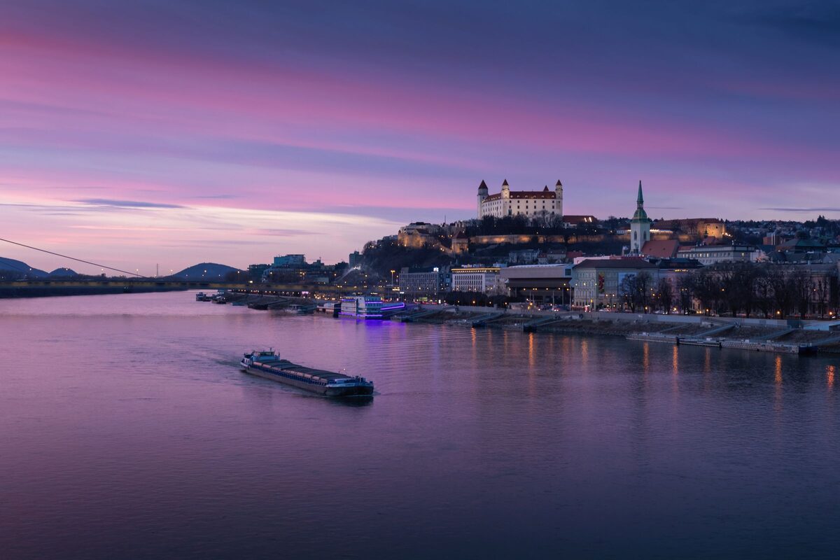 Bratislava Castle lit up on its hill above the Danube at dusk, with a barge on the river