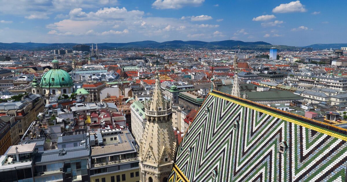 View over Vienna's rooftops from St. Stephen's Cathedral, with the cathedral's zigzag tiled roof and Gothic spire in front and the Vienna Woods on the horizon