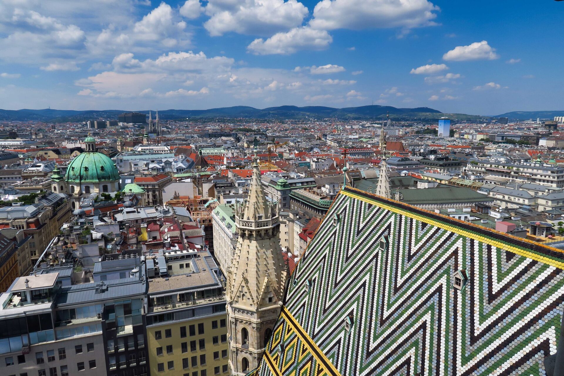 View over Vienna's rooftops from St. Stephen's Cathedral, with the cathedral's zigzag tiled roof and Gothic spire in front and the Vienna Woods on the horizon