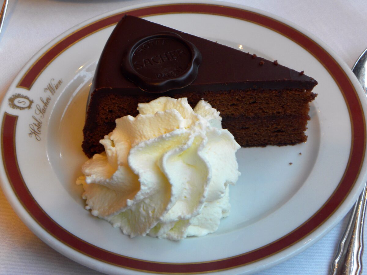 A slice of Sachertorte with whipped cream on a Hotel Sacher plate, with the chocolate seal on top