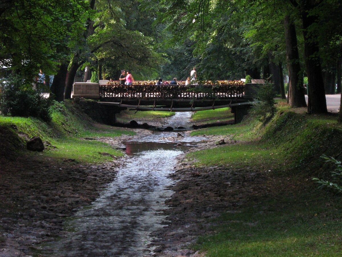 The Bridge of Love seen from the bed of the Vrnjačka river, its railing thick with padlocks, under the shade of tall park trees.