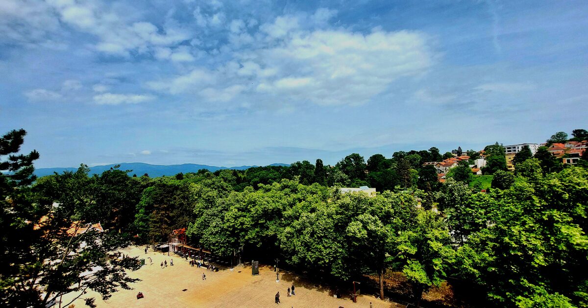 The paved square of the central park in Vrnjačka Banja seen from above, framed by thick green treetops with the Goč range on the horizon.