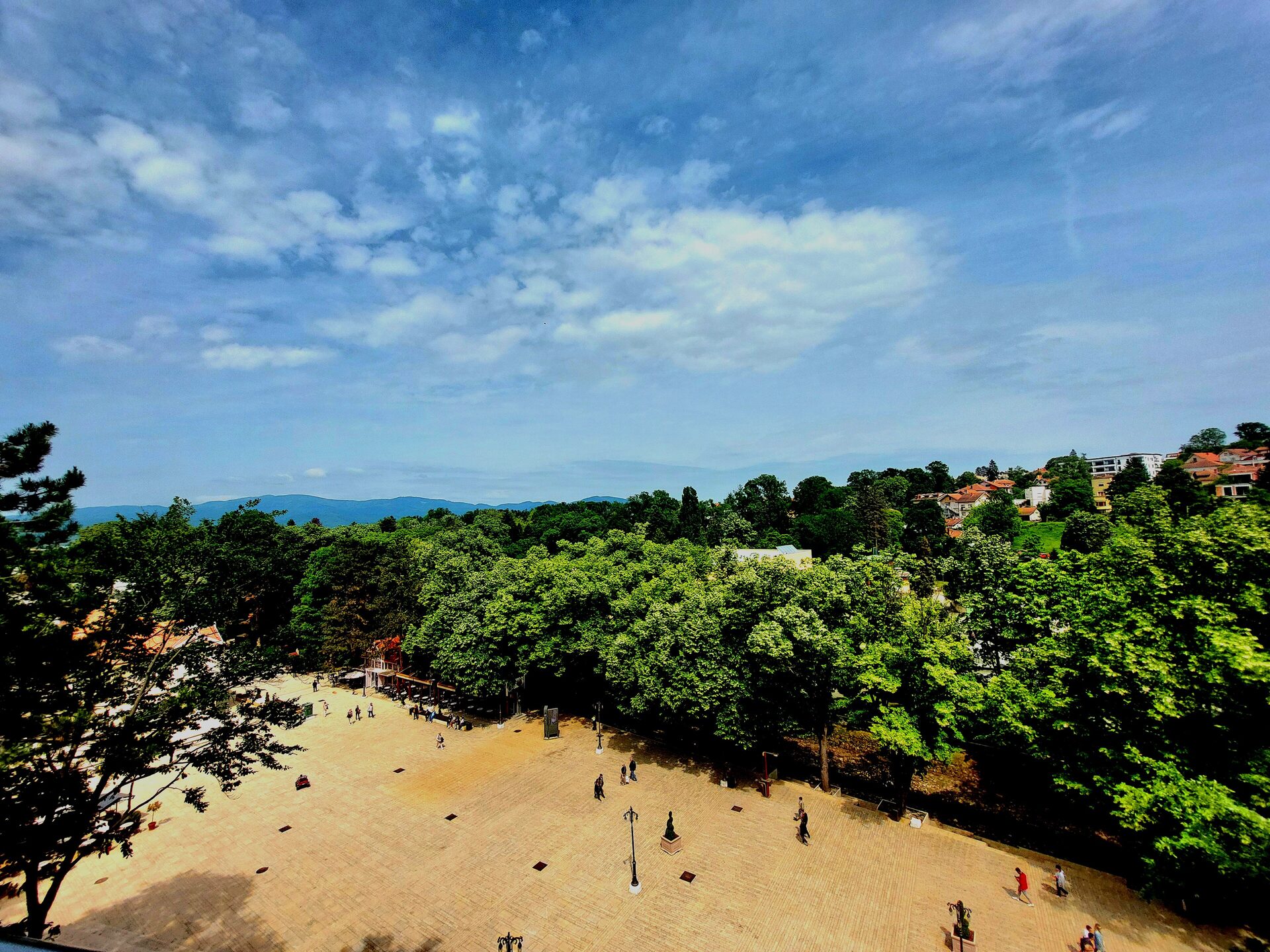 The paved square of the central park in Vrnjačka Banja seen from above, framed by thick green treetops with the Goč range on the horizon.