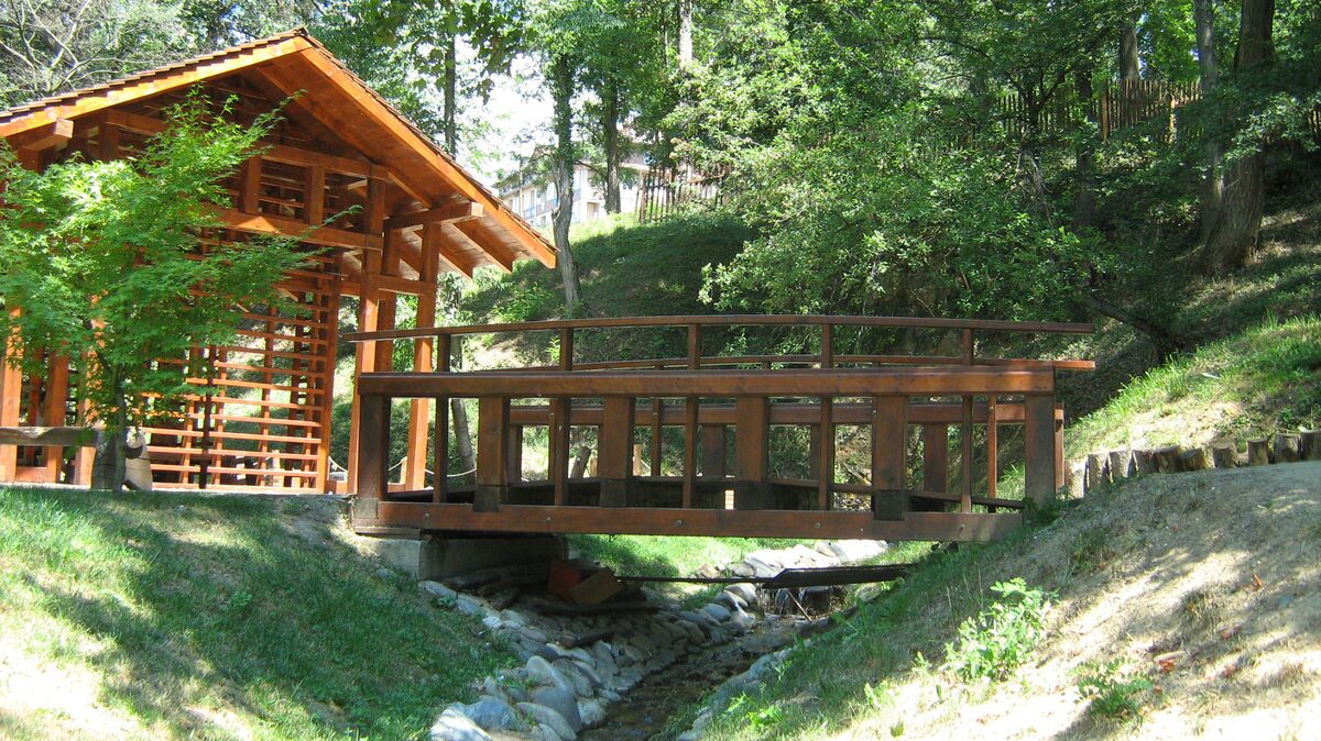 A wooden pavilion and a wooden footbridge over a stony stream in the Japanese Garden, surrounded by grassy banks and leafy trees.