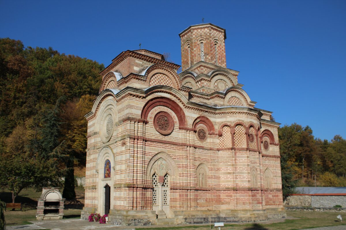 The church of Kalenić monastery in striped stone and red brick with carved rose windows and a dome, against a clear blue sky and autumn woods.