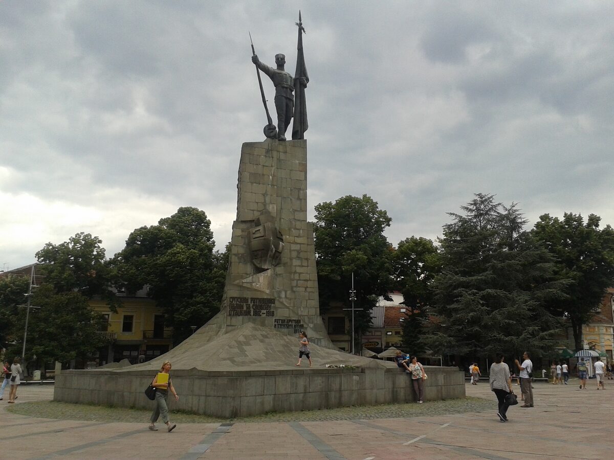 The Monument to the Serbian Warrior rising from the paved round square in central Kraljevo, with trees and low buildings around it under a cloudy sky.