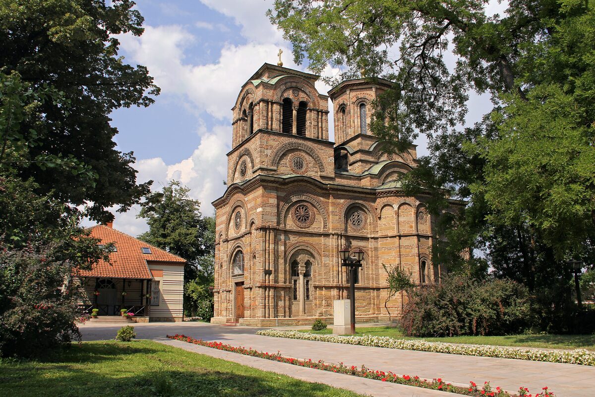 Lazarica church in Kruševac, built in banded stone and brick with rose windows and a bell tower, framed by trees and a path lined with flowers.