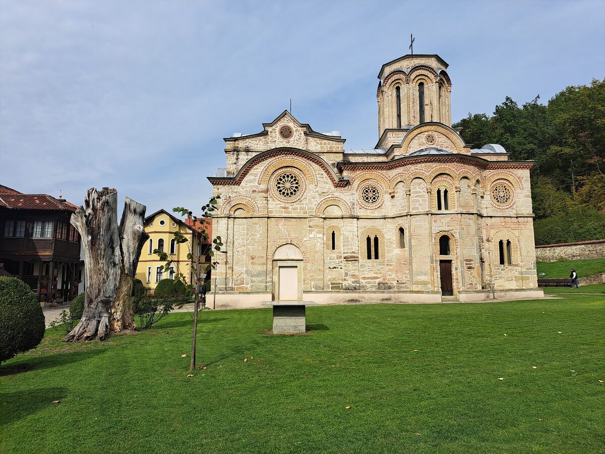 The stone Church of the Dormition at Ljubostinja monastery with rose windows and a single dome, across a green lawn beside an old tree stump and the monastery lodgings.