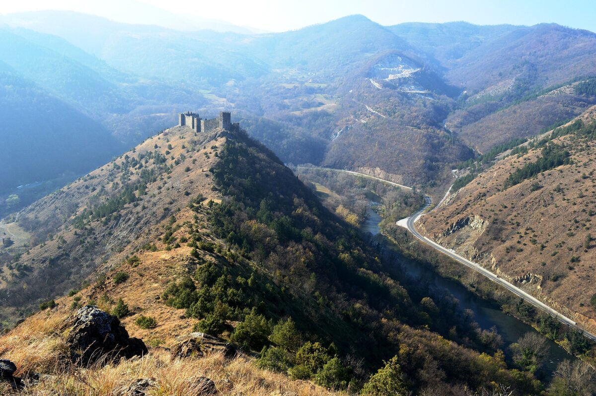 The ruined towers of Maglič fortress on a steep ridge above the Ibar gorge, with the river and a road winding along the valley floor below.
