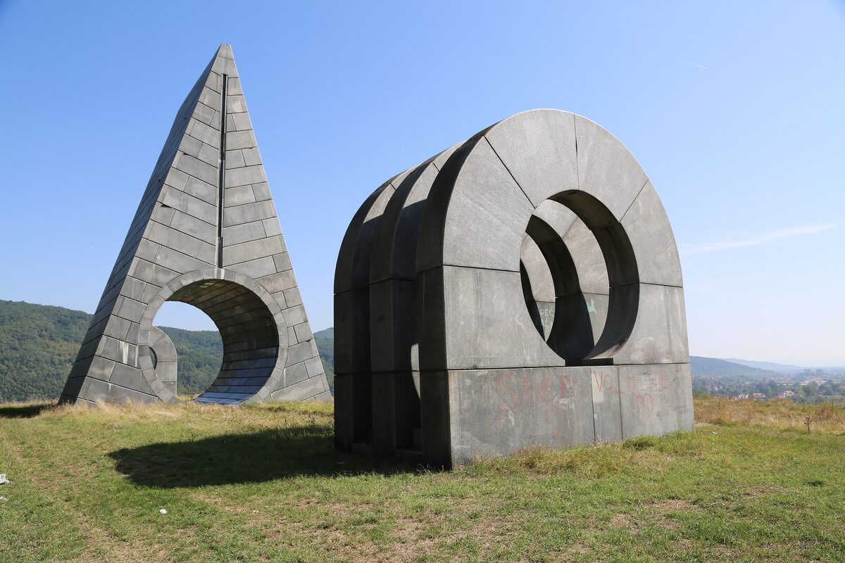 Two stone sculptures by Bogdan Bogdanović at Popina memorial park, a pointed pyramid and a block pierced by stepped circular openings, on a grassy hilltop.