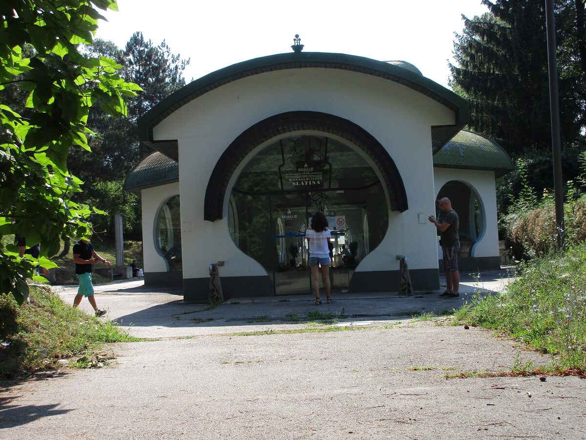 The white Slatina spring pavilion with its green rounded roof and a large arched glass entrance, set among trees in the park.