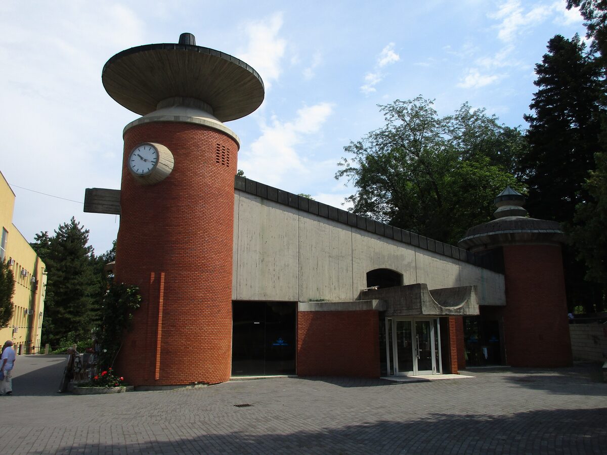 The Topla voda spring pavilion, a brick and concrete building with a round clock tower capped by a mushroom-shaped roof, under a summer sky.