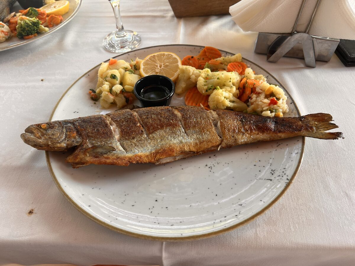 A whole grilled trout with scored, crisp skin on a white plate, served with lemon, steamed vegetables and a small dish of oil.