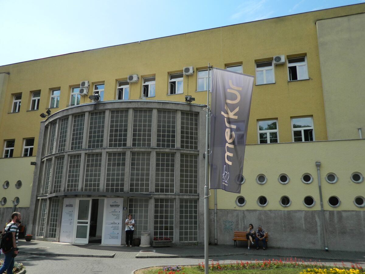 The yellow interwar bath building of the Merkur spa, with a curved glass-block entrance, round porthole windows and a Merkur banner out front.