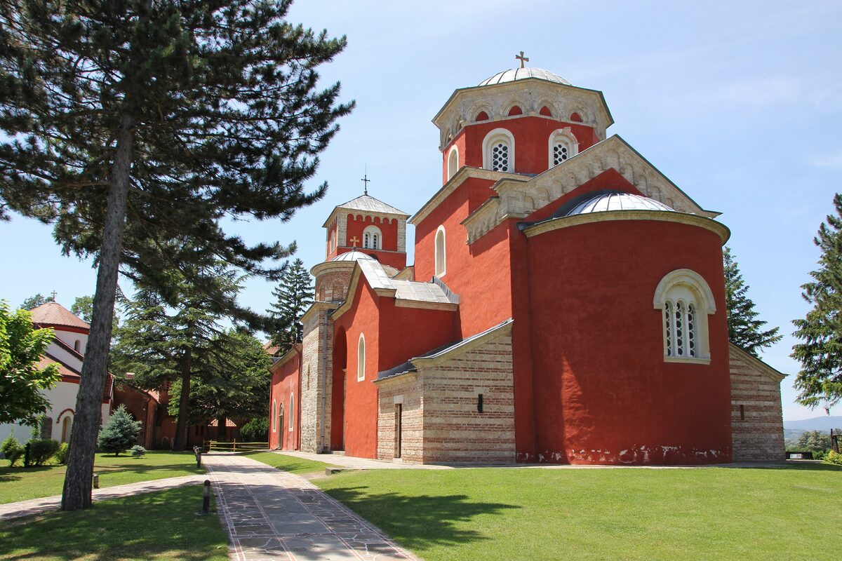 The red-painted Church of the Ascension at Žiča monastery with its dome and west tower, beside a tall pine and a paved path across the lawn.