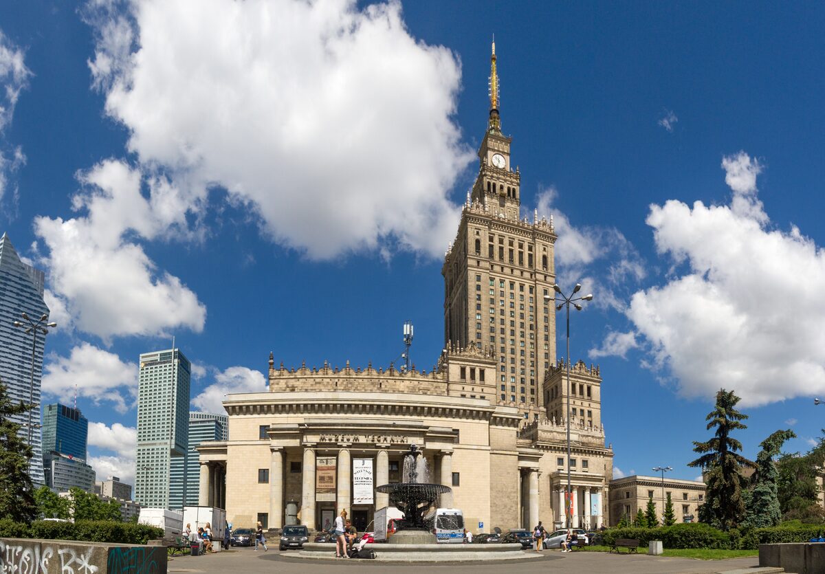 The Palace of Culture and Science, a tall Stalinist tower with a spire, above its colonnaded entrance and a fountain, under a blue sky with clouds