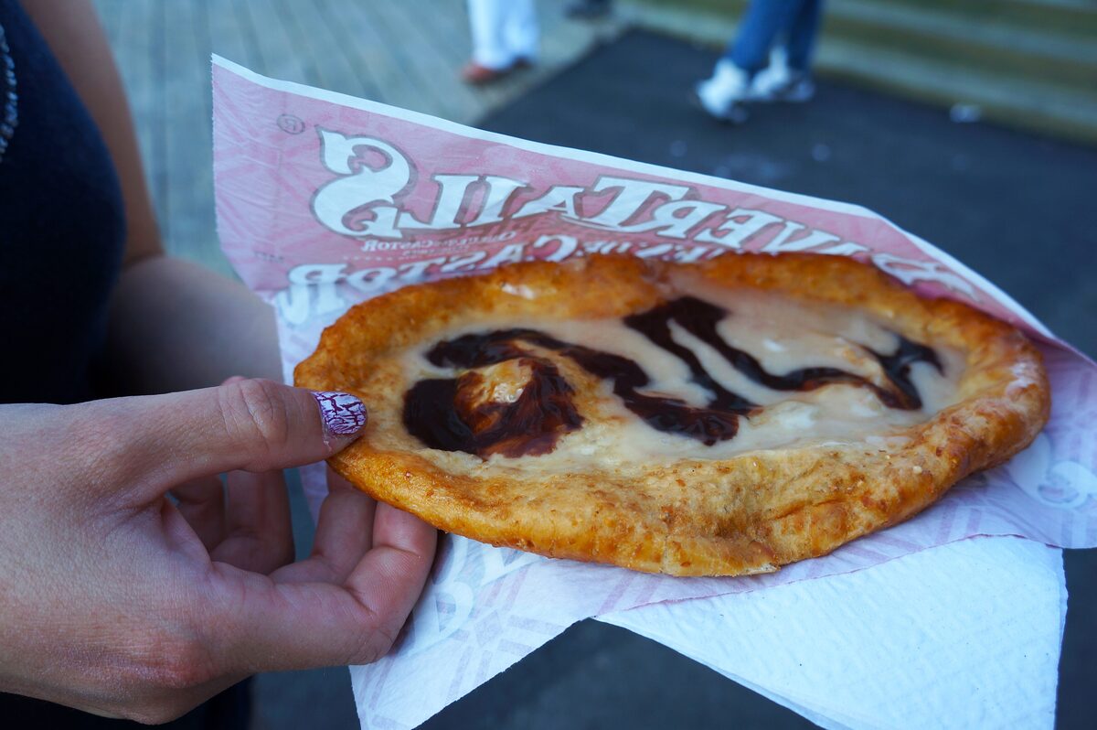 A hand holding a flat fried BeaverTail pastry with chocolate and white topping, on a BeaverTails paper sleeve
