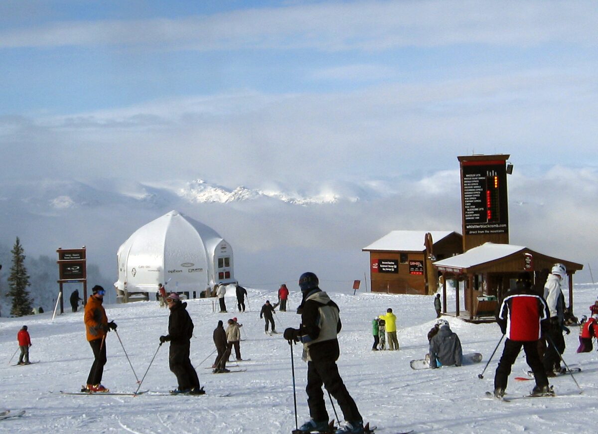 Skiers and snowboarders on the snow in front of the Roundhouse Lodge area, with clouds in the valley below