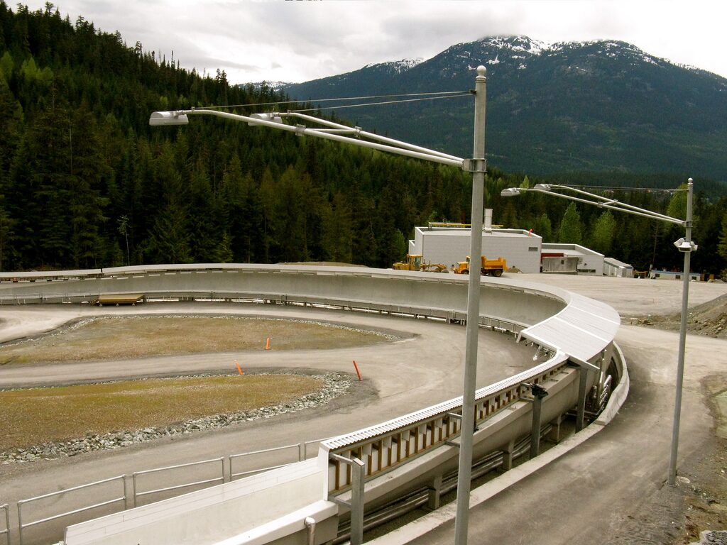 A curved bend of the Whistler Sliding Centre track below forested mountains, seen in summer without snow or ice