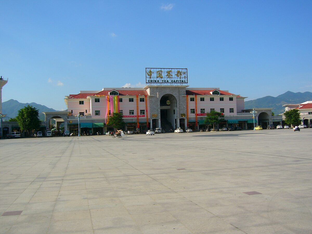 The China Tea Capital trading complex in Anxi County, a pink and white building with a stone gateway beyond a wide empty square, with hills behind