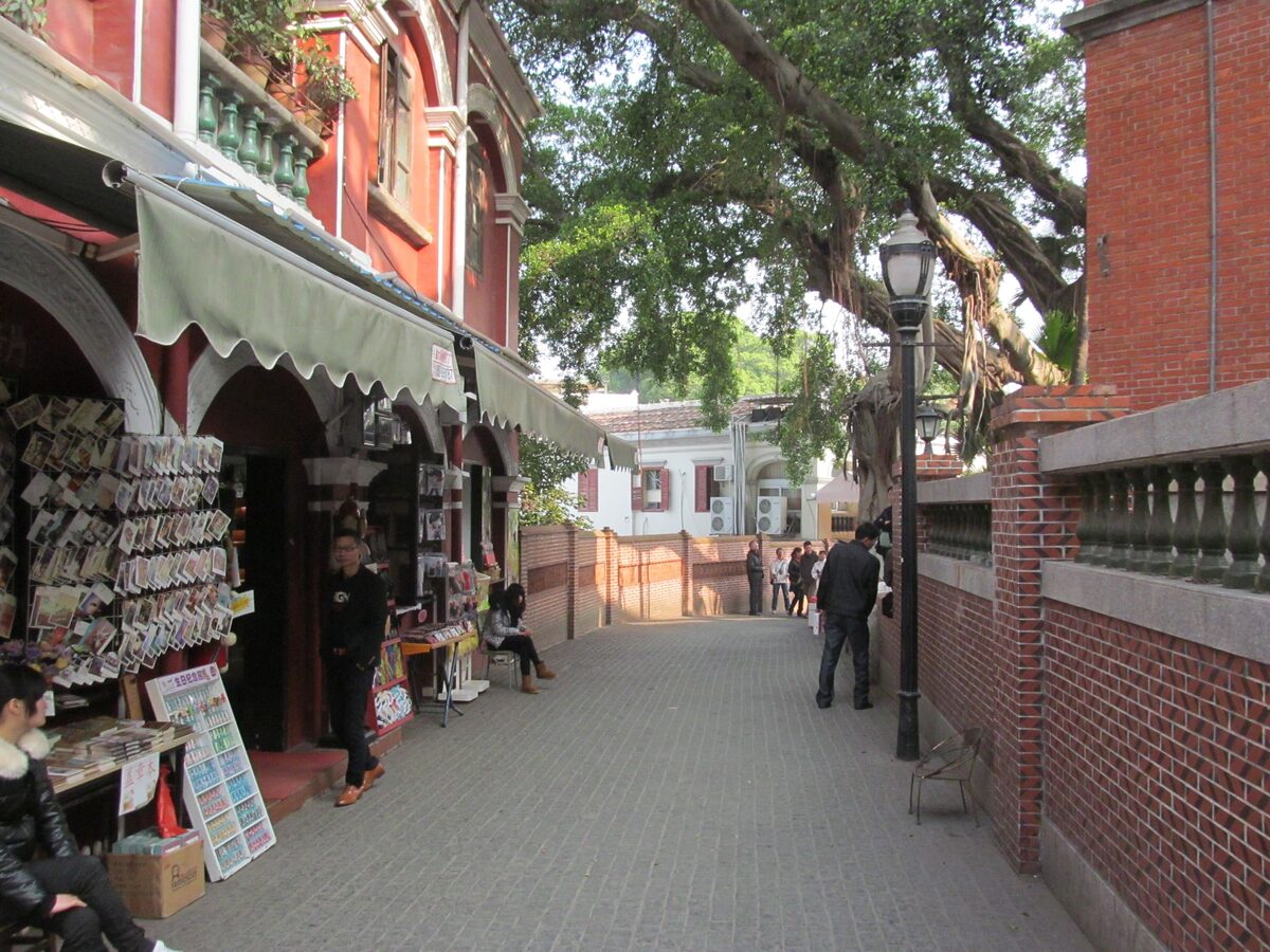A paved Gulangyu lane with souvenir stalls on one side and a red-brick villa wall on the other