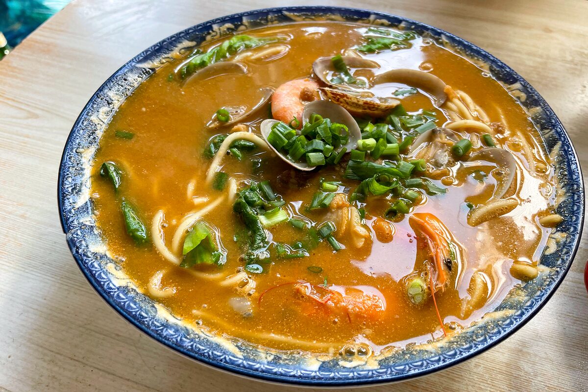A bowl of seafood shacha noodles in orange broth with shrimp, clams and spring onion