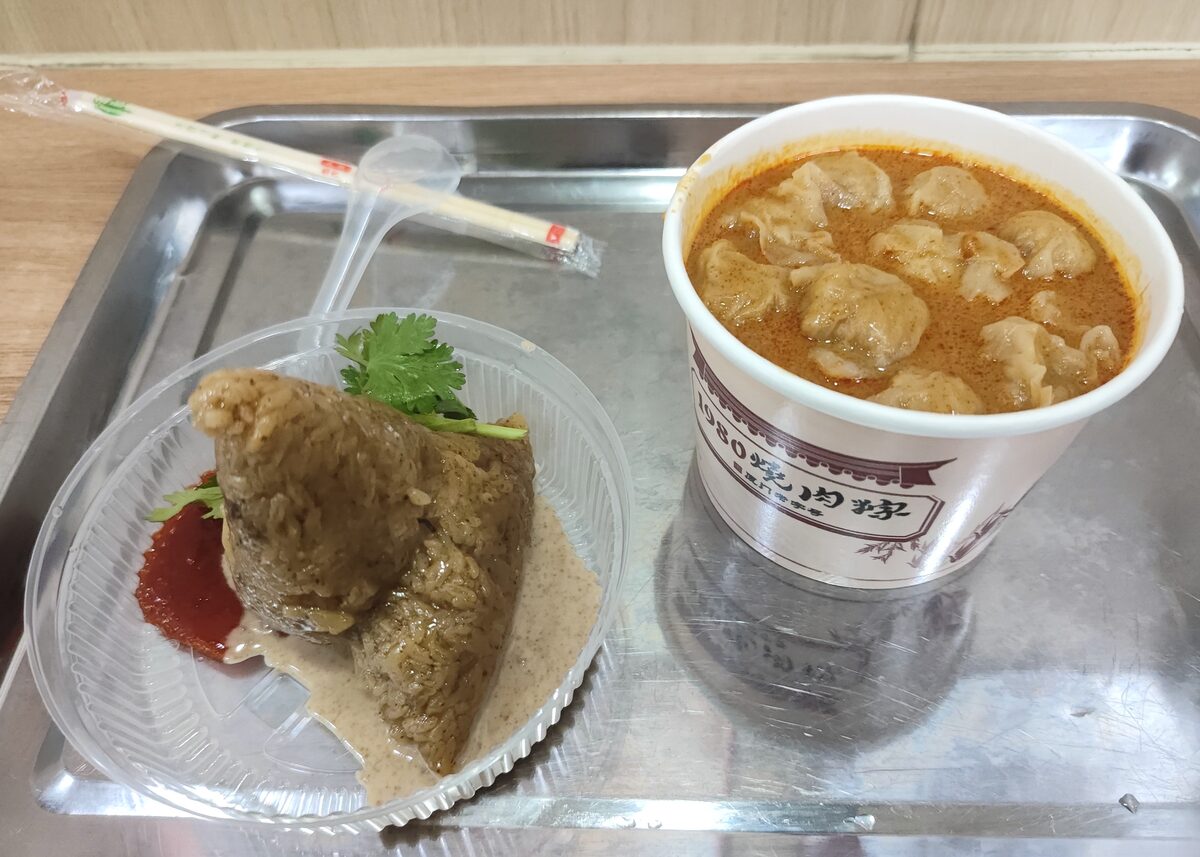 A glutinous rice dumpling with red chili sauce on a tray, beside a paper cup of dumplings in orange broth
