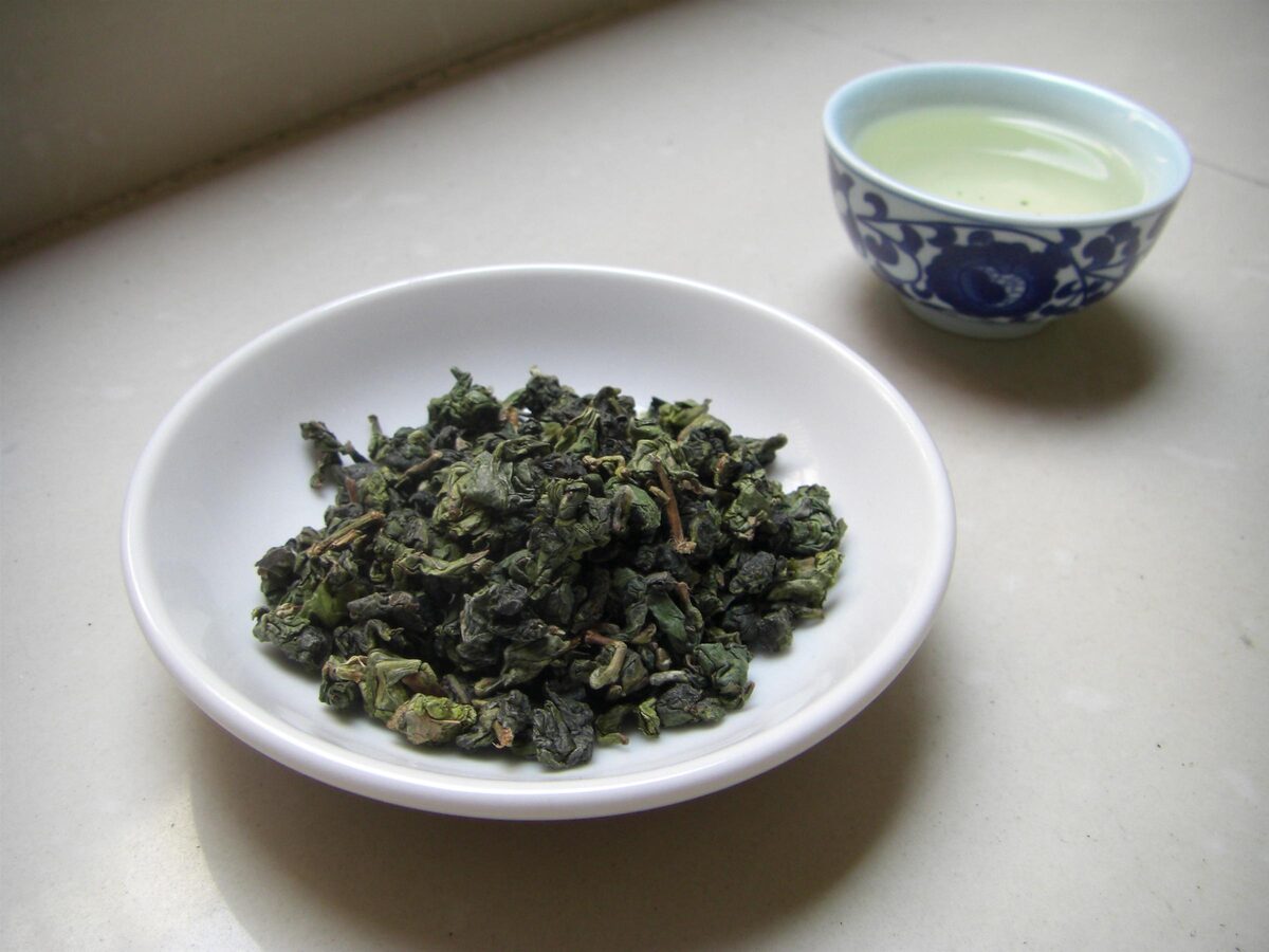 A dish of rolled Tieguanyin oolong leaves beside a cup of pale green tea