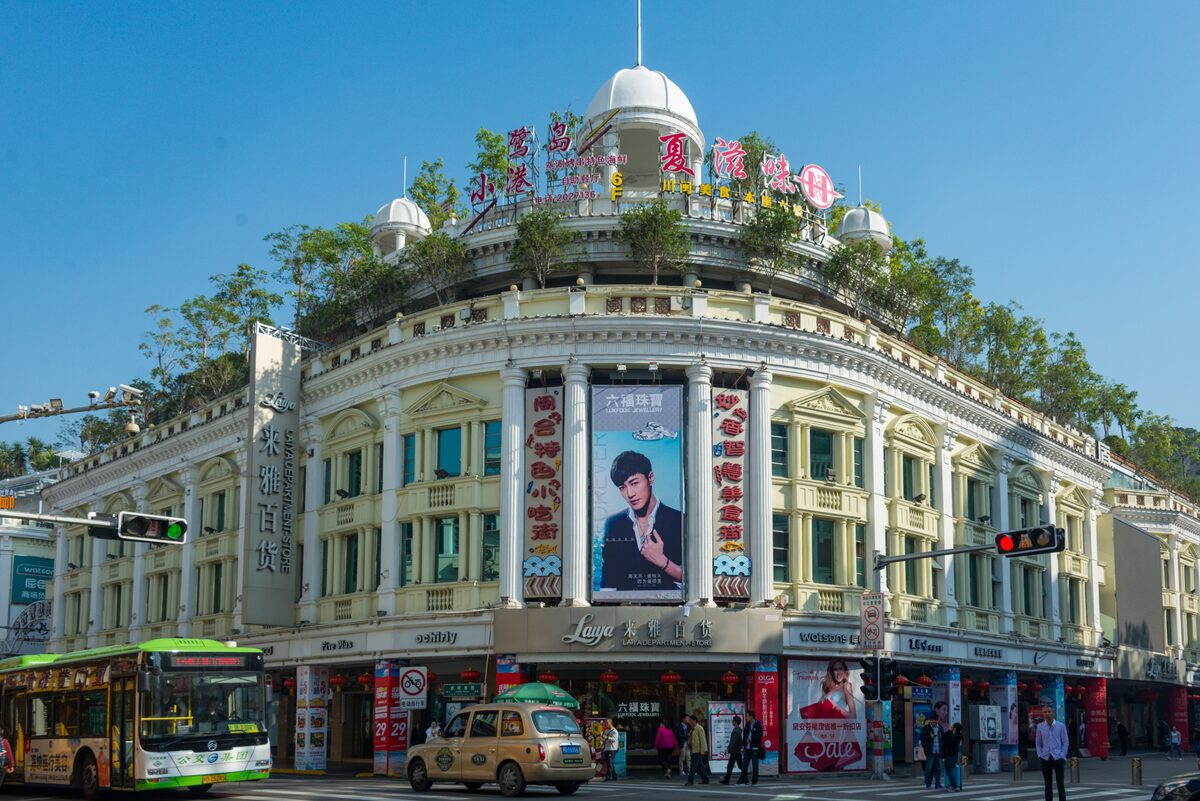 A cream-colored colonial-style corner building with a small dome and shops at street level on Zhongshan Road, with a bus and a taxi passing
