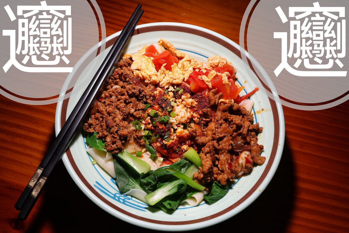 A plate of belt-wide biangbiang noodles topped with spiced minced meat, greens and tomato, with the complex character for biang printed beside it