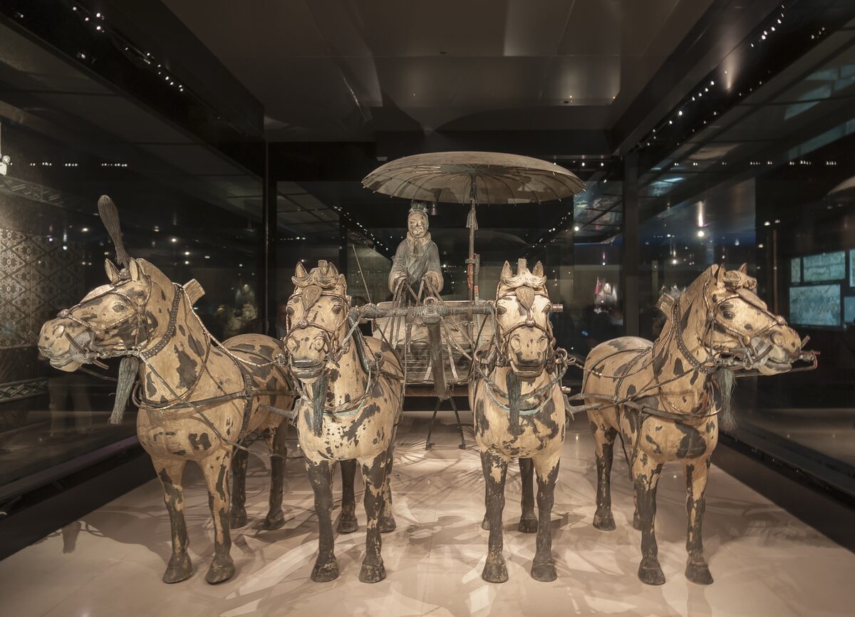 A bronze chariot with four bronze horses and a seated driver under a canopy, displayed in a glass case