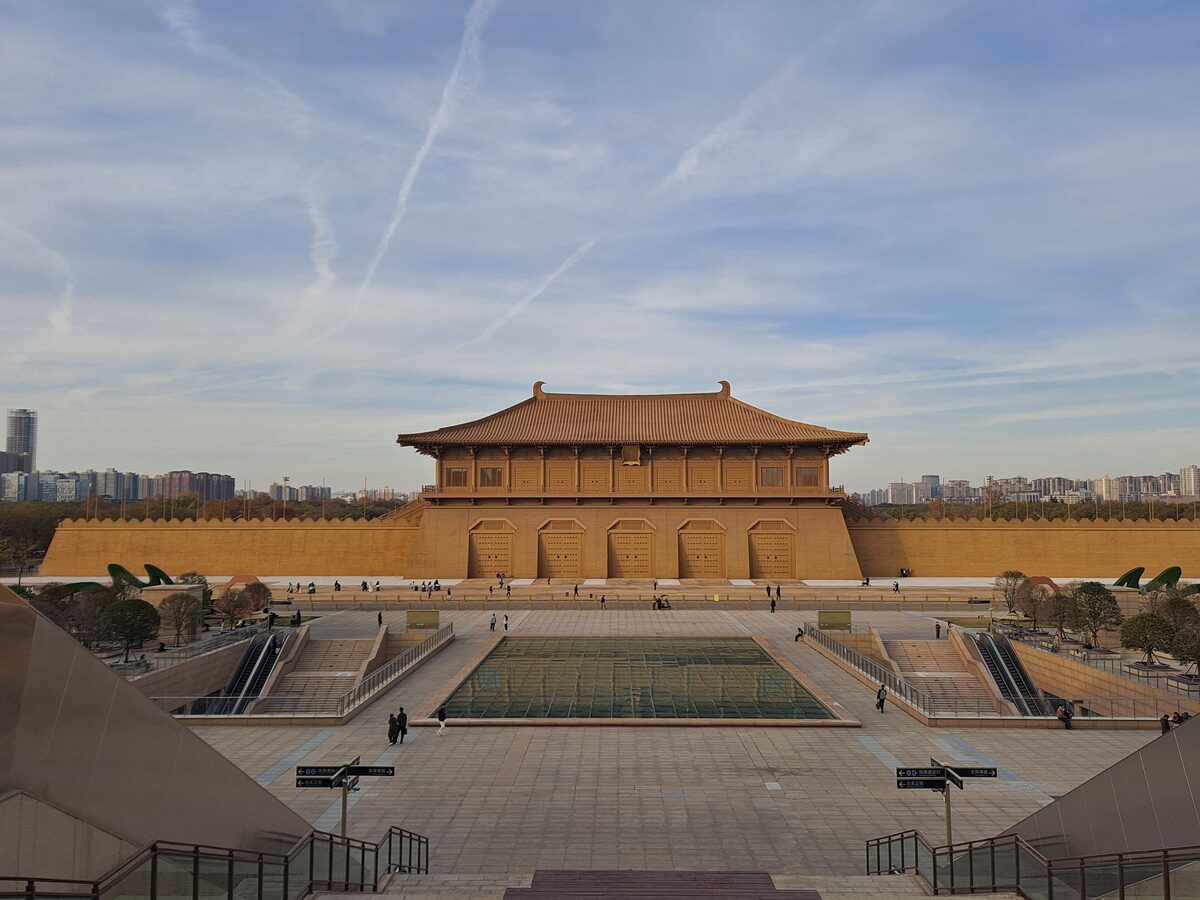 The rebuilt Danfeng Gate of Daming Palace, a wooden gate tower on a rammed-earth base, seen across a wide plaza under a sky streaked with contrails
